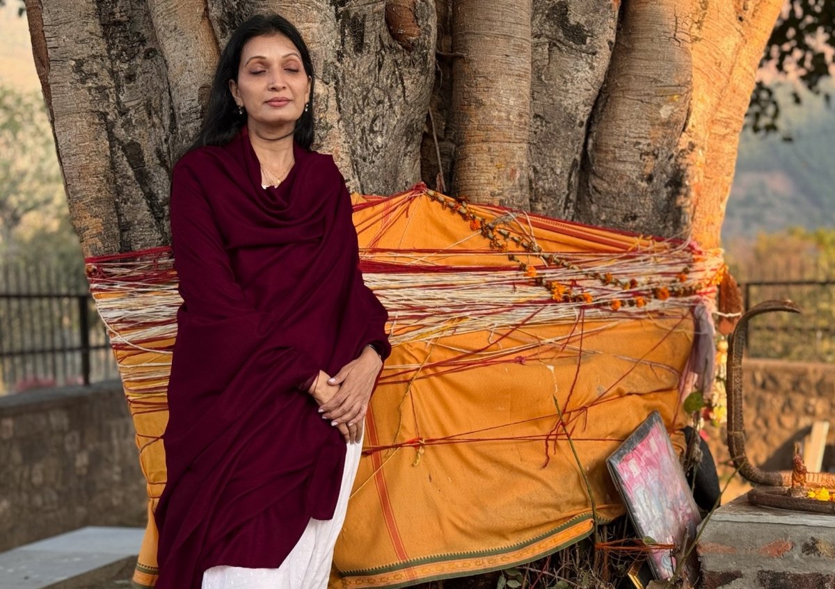 Woman in a meditation state wrapped in a large deep red pashmina shawl standing in front of a Indian temple tree.