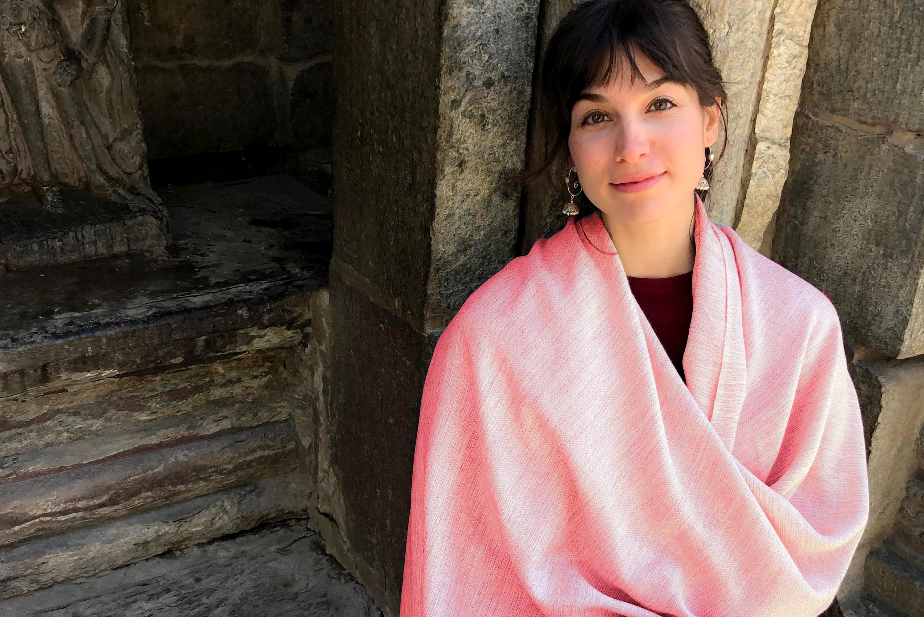 A person wrapped in a burnt orange merino wool meditation shawl with fringes, standing near stone stairs.