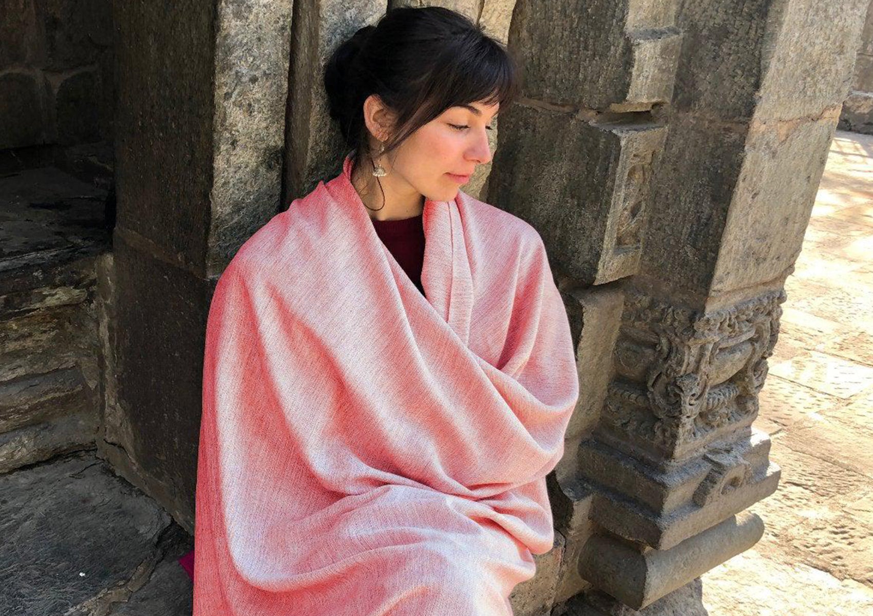 A person wrapped in a burnt orange merino wool meditation shawl with fringes, standing near stone stairs.