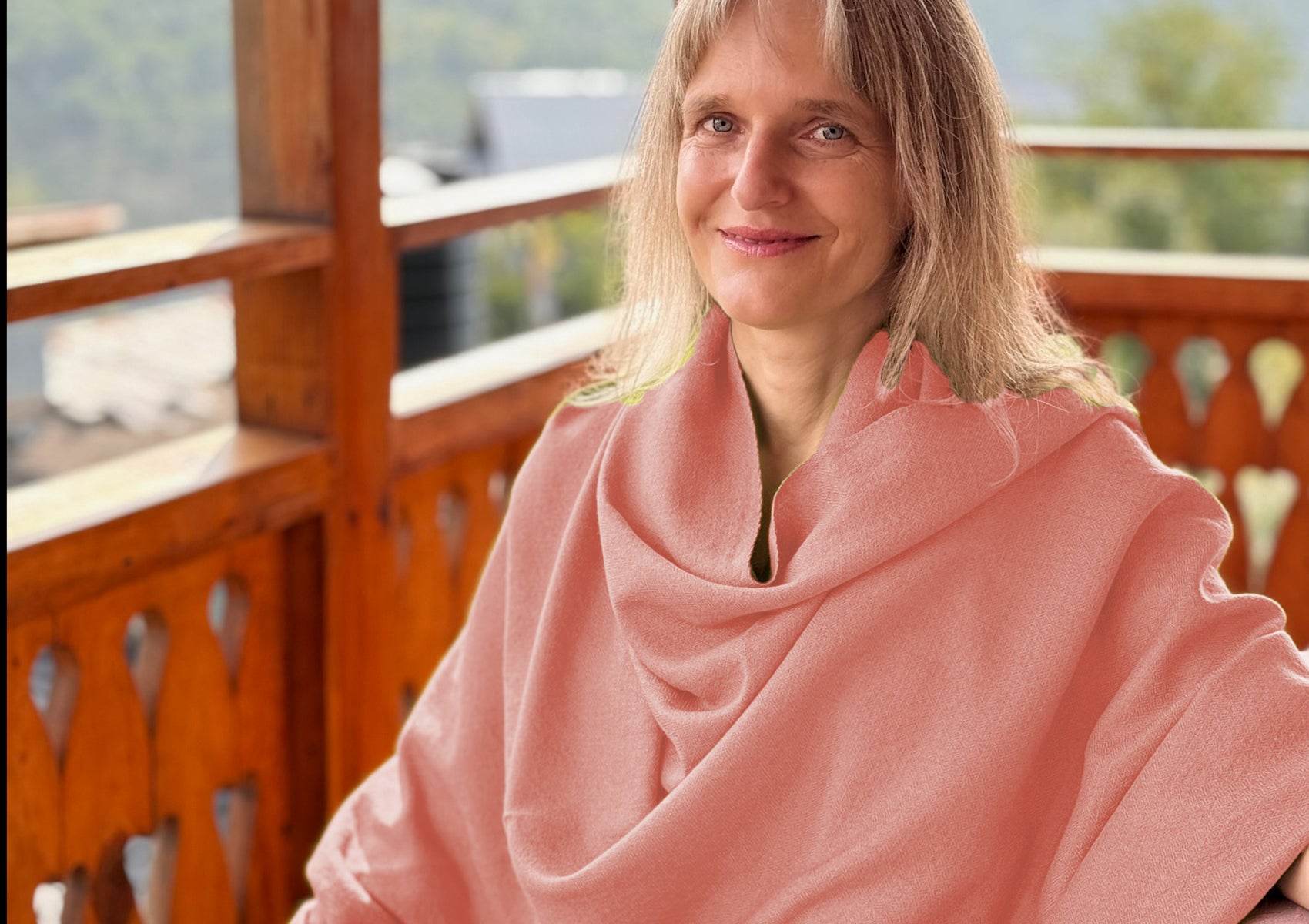 Woman wearing peach meditation shawl, seated on a wooden balcony in serene pose.