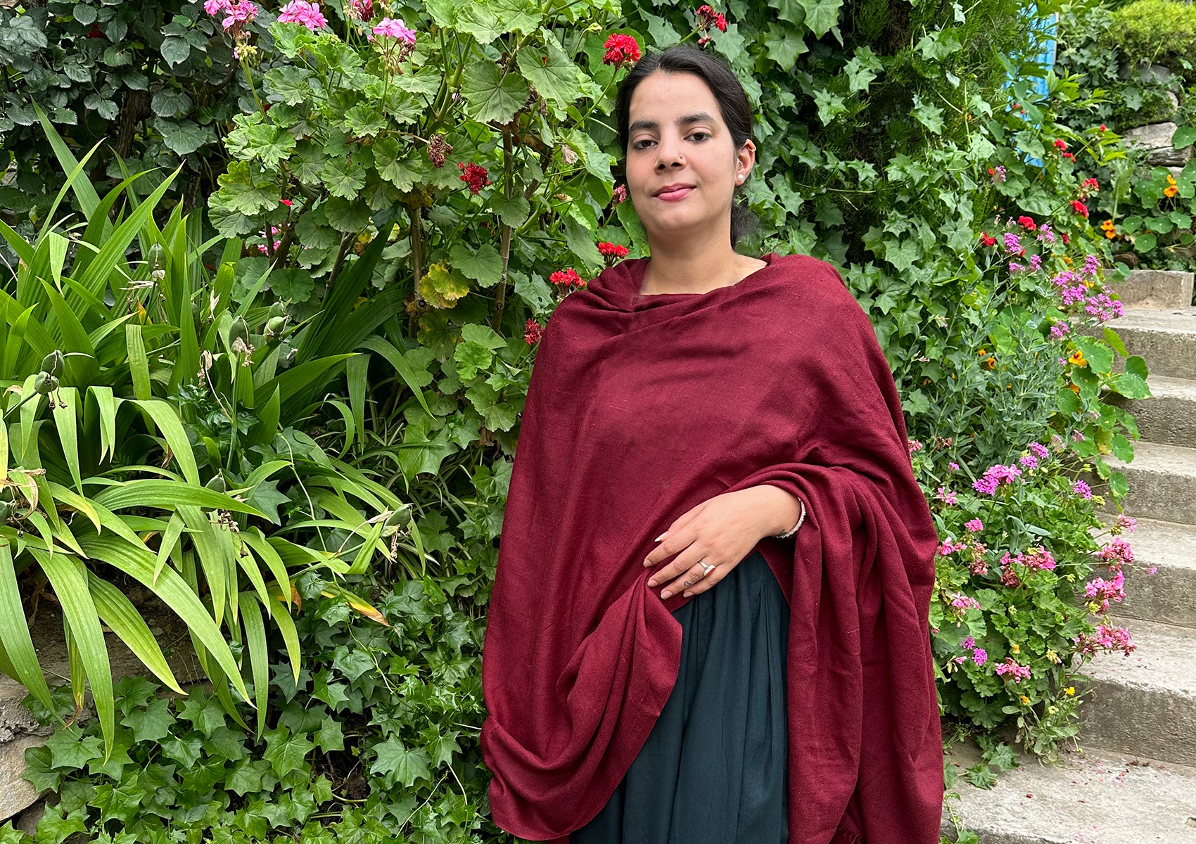 Woman wearing a burgundy meditation shawl, standing in a lush garden filled with vibrant flowers and greenery.