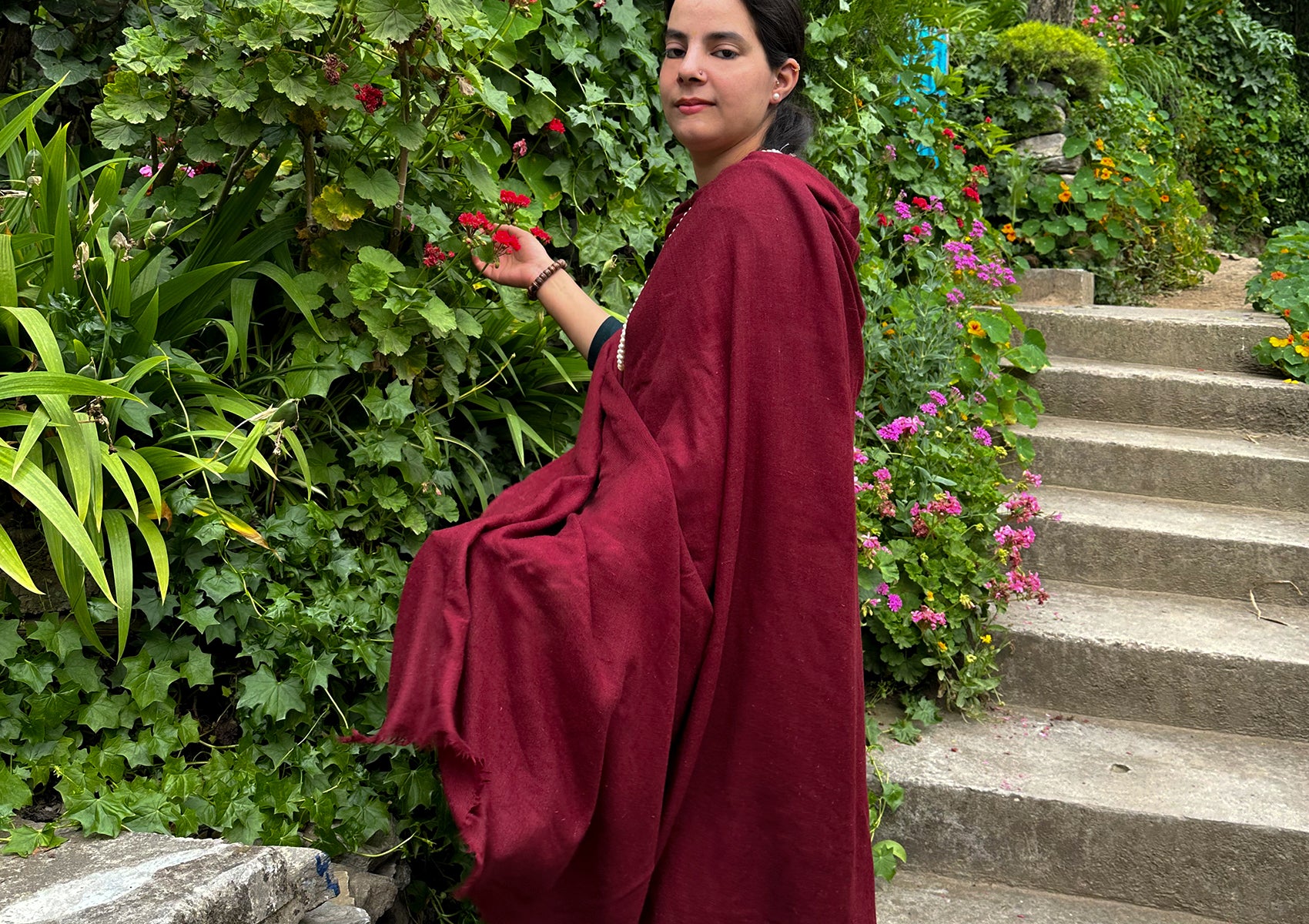 Woman in a burgundy meditation shawl, standing beside vibrant garden steps, holding a red flower.
