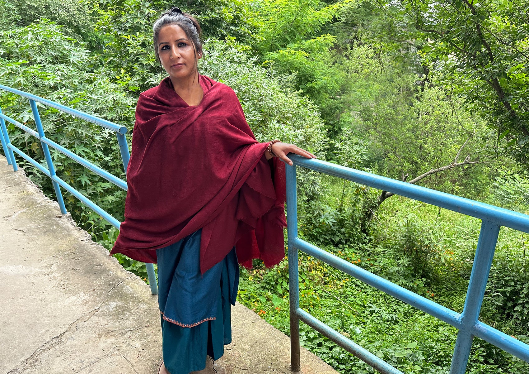 Model wearing a burgundy meditation shawl standing near a blue railing, surrounded by lush greenery.