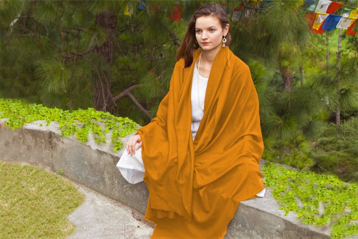 Woman sitting in a garden, wrapped in a soft orange shawl, with prayer flags and greenery in the background.
