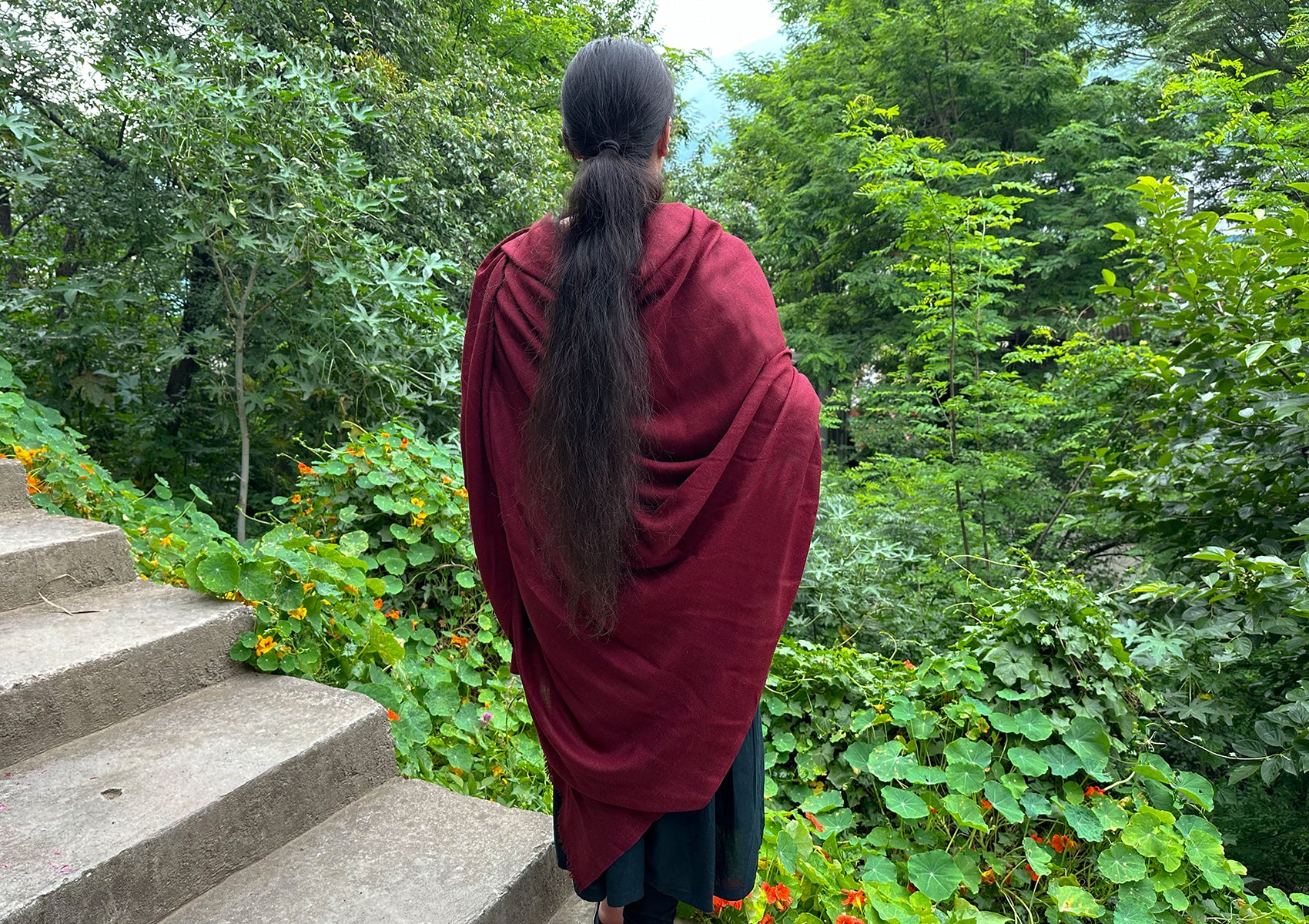 Woman in a red meditation shawl surrounded by lush green garden foliage, enjoying nature.