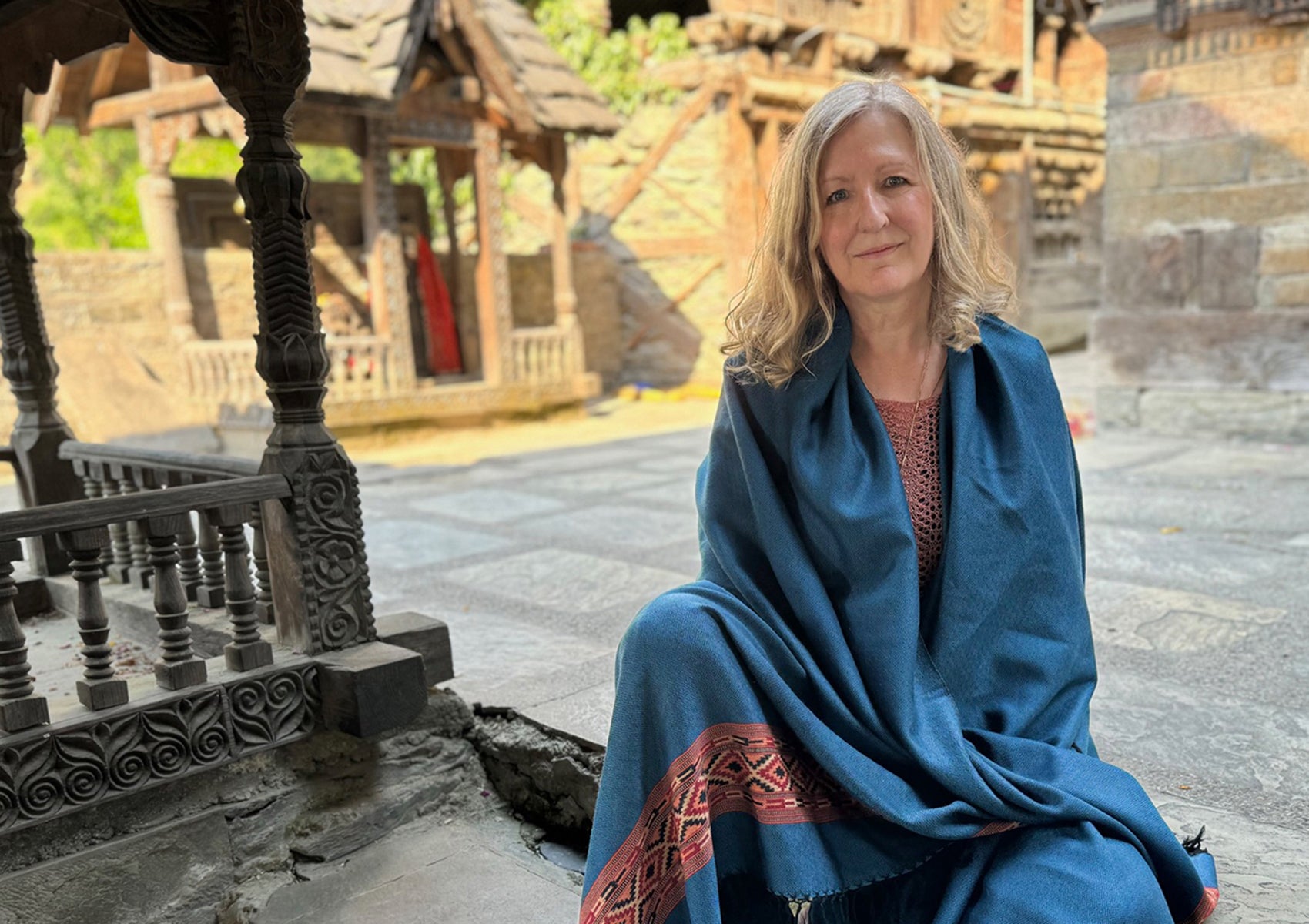 Woman sitting in temple courtyard wrapped in DEVA denim blue merino wool shawl with ethnic border.