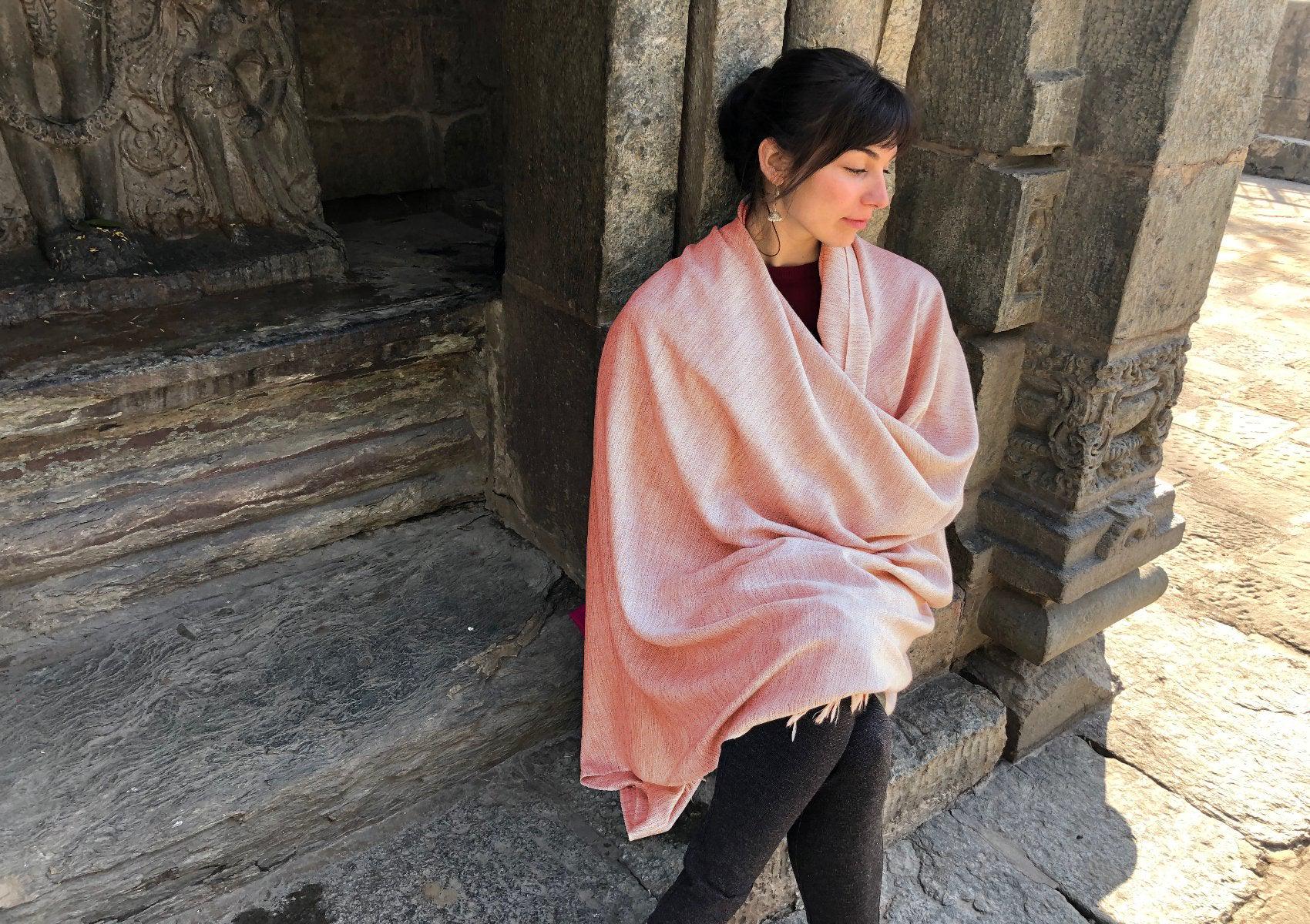 Woman wrapped in a burnt orange shawl standing next to an ancient stone background, exuding a calm vibe.