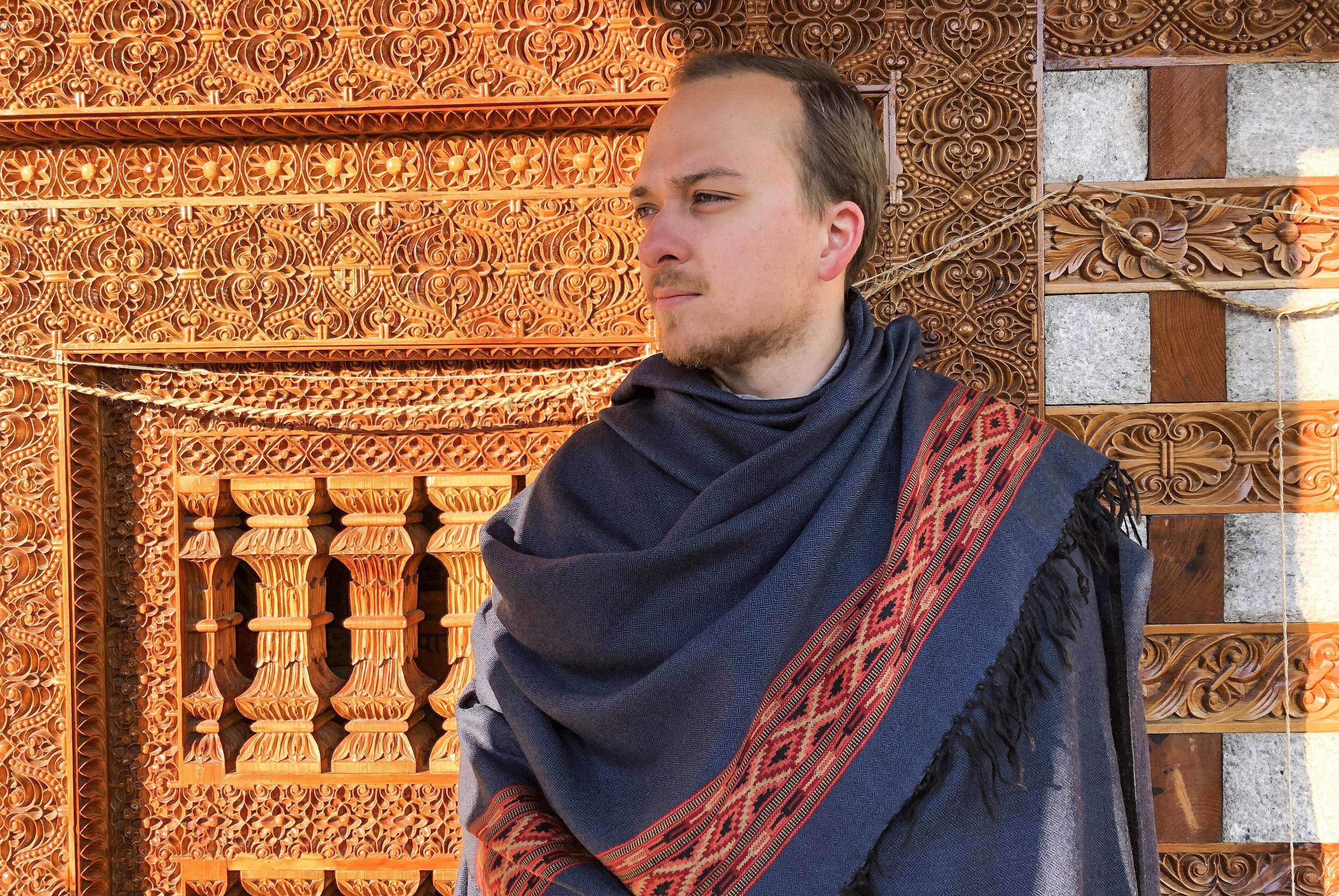 Man wearing DEVA Steel Blue meditation shawl with traditional ethnic border in front of carved wooden temple wall