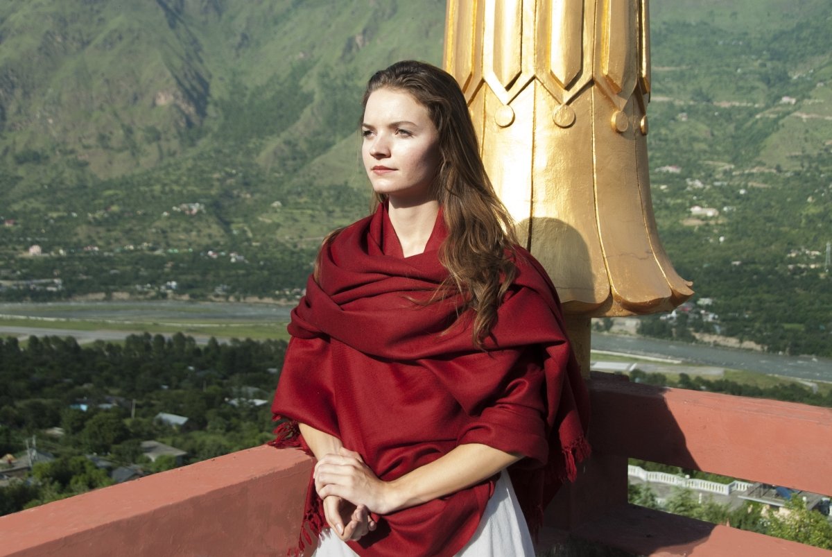 A woman smiling, wrapped in a red shawl, with golden stupa and mountains in the background.