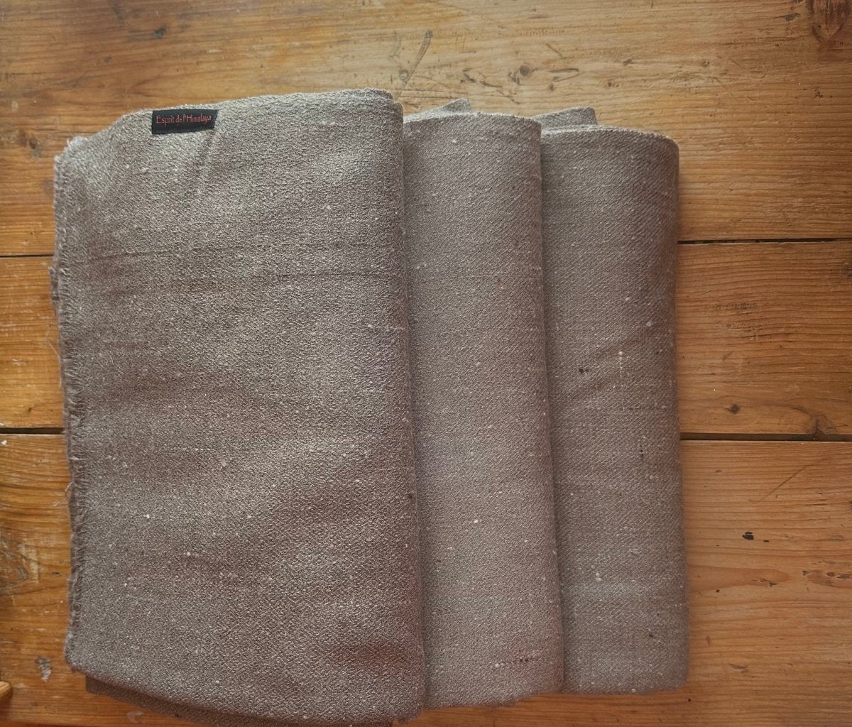 three dark beige meditation shawls folded on a wooden floor.