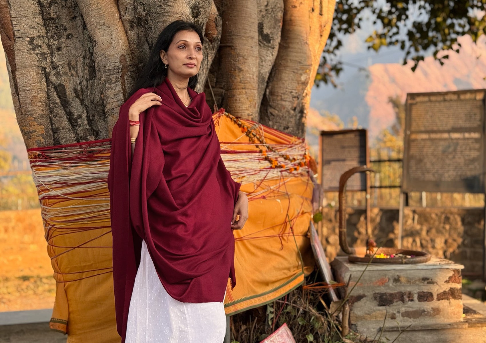 Woman wearing a handwoven burgundy pashmina meditation shawl draped gracefully beside a sacred Himalayan tree at golden hour.