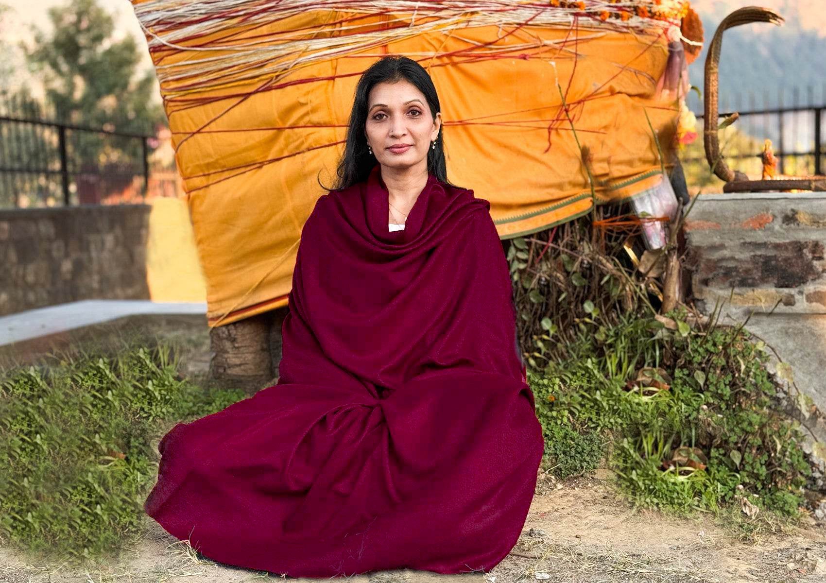 Woman seated in meditation wrapped in a deep burgundy handwoven pashmina shawl near a sacred Himalayan tree at sunset.