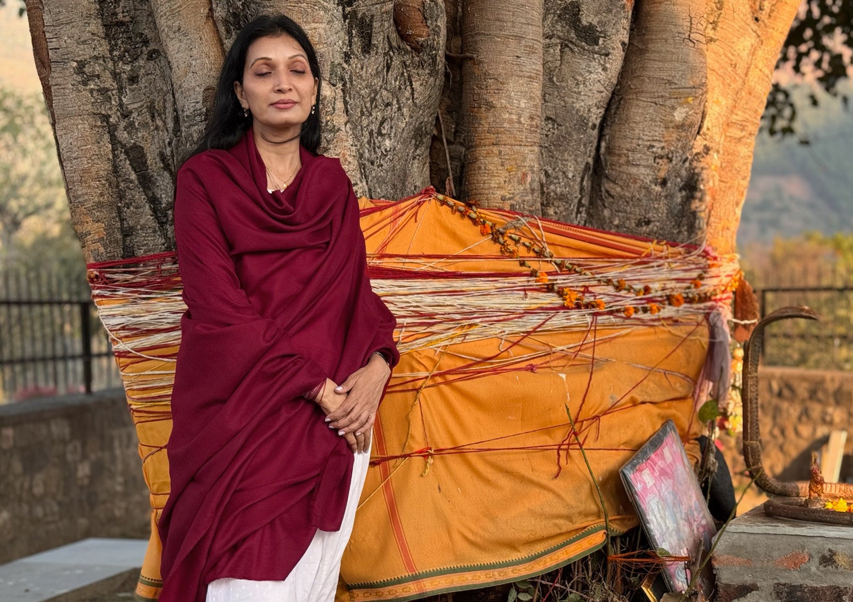Woman wrapped in a deep burgundy handwoven pashmina shawl in a peaceful meditative pose near a sacred tree in the Himalayas.