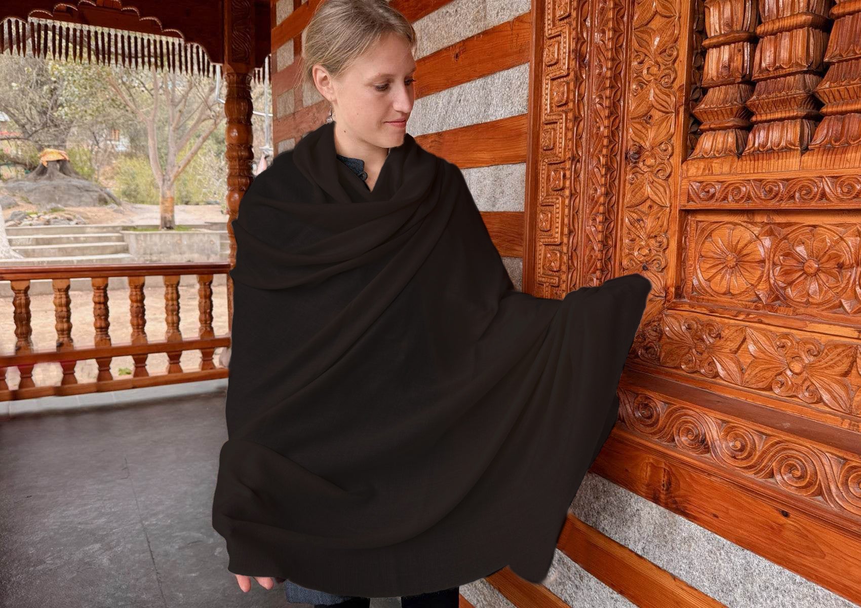 A women wrapped in a Bodhi chocolate brown large pashmina shawl standing near a wooden sculpted himalayan temple.