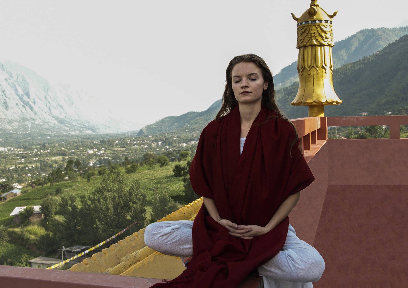 Woman over a rooftop, draped in a burgundy shawl, with a golden temple ornament and lush mountain view.