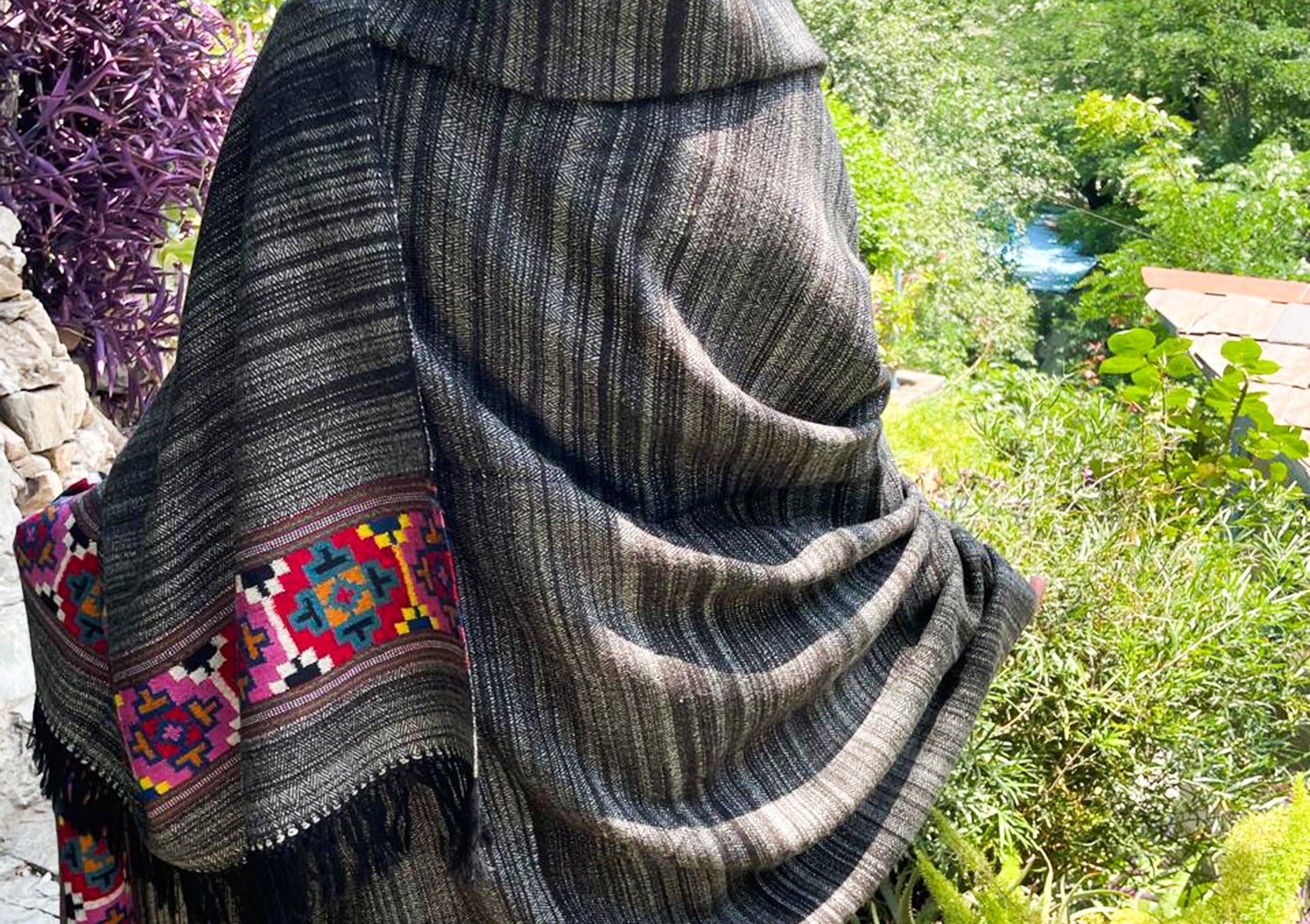 Woman draped in a thick grey yak wool shawl (Girish Gulab) with a peeking vibrant hand-woven colorful border, viewed from the side in a garden setting.