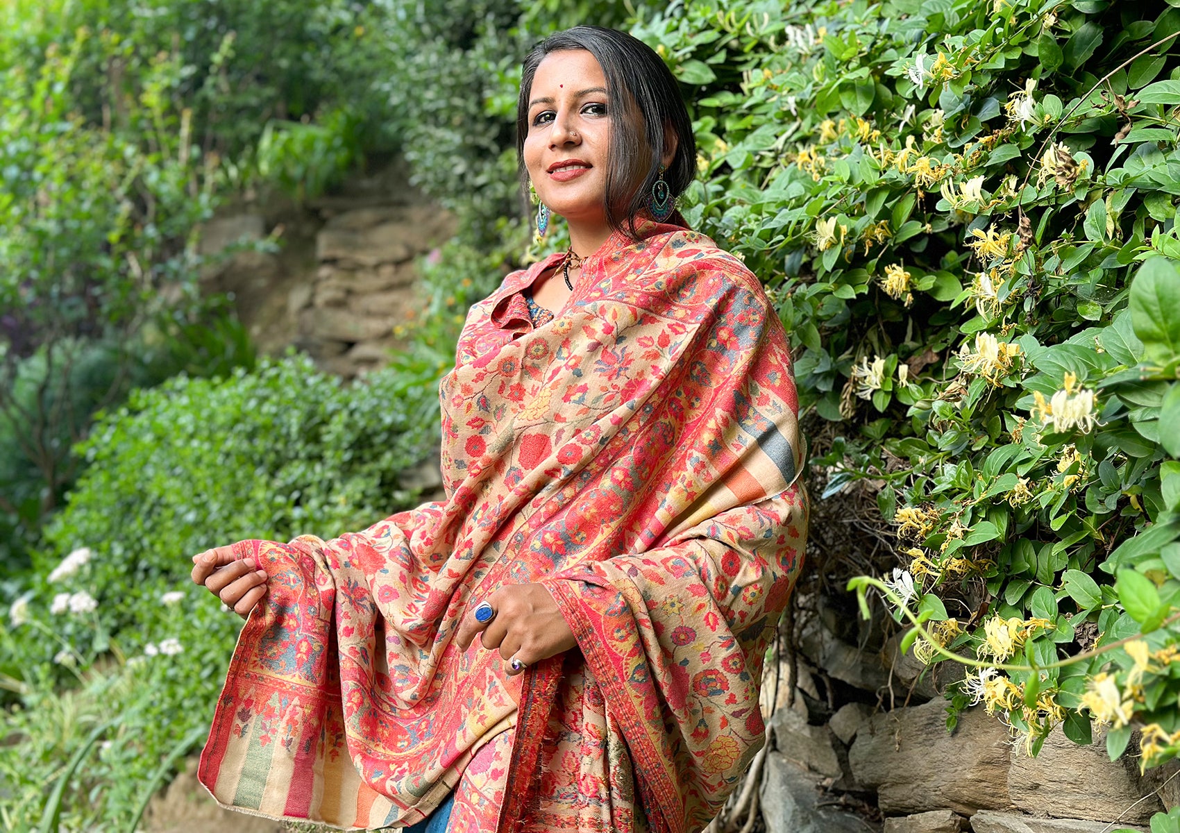 Person elegantly wrapped in a beige Mohani Kani shawl adorned with vibrant floral patterns, standing against a lush garden with blooming yellow flowers.