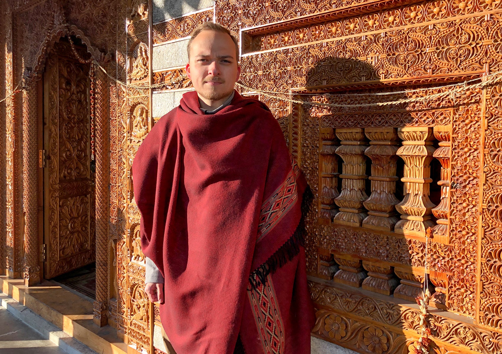 Man wearing DEVA dark red merino wool meditation shawl with ethnic Kinnauri border in front of carved temple wall.