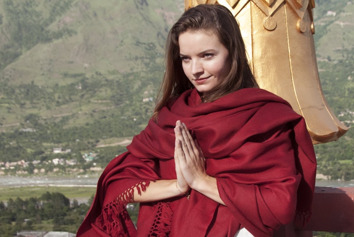Side view of a person meditating against a temple wall, dressed in a red meditation shawl.